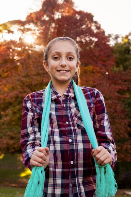 Young girl smiling and enjoying a sunny autumn day in the park. She is wearing a plaid shirt and holding a light blue scarf. The background features colorful autumn leaves and sunlight filtering through the trees. Perfect for use in advertisements, seasonal promotions, family-oriented content, and outdoor activity themes.