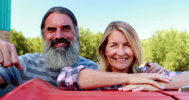 A middle-aged Caucasian couple smiles joyfully while leaning on a red vehicle, with copy space. Their cheerful expressions and casual attire suggest a moment of leisure or a road trip adventure.