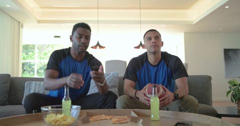 Diverse friends watching game on sofa holding football and pizza during casual game night