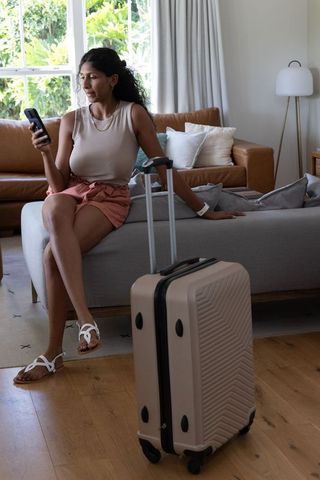 African american woman sitting on sofa checking smartphone beside beige carry-on luggage