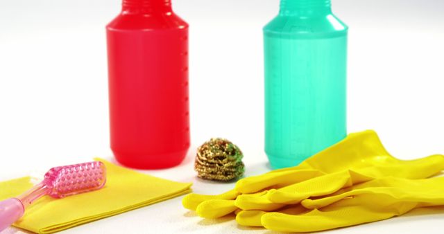 Colorful cleaning supplies on white surface, featuring red and blue detergent bottles, yellow gloves, a scrubber, a pink brush, and a yellow cloth. Ideal for use in articles and publications about household chores, hygiene, cleaning tips, and maintenance. Suitable for home improvement blogs, cleaning service websites, and instructional content on cleaning methods.