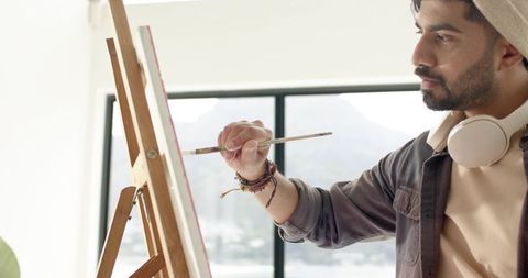 Indian artist painting on canvas at easel in bright studio wearing beanie and headphones