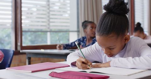 Focused child writing in classroom by window with blinds
