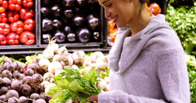 Woman wearing casual hoodie, smiling while holding bunch of fresh green vegetables at grocery store. Perfect for promoting healthy lifestyle, grocery shopping, fresh produce markets, or healthy eating habits.