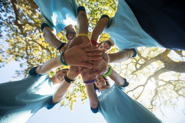 This image shows a group of volunteers forming a hand stack, symbolizing unity and teamwork. The low angle view and outdoor park setting emphasize the collaborative spirit and connection with nature. Ideal for use in promotional materials for community events, charity organizations, teamwork and collaboration themes, or social work initiatives.