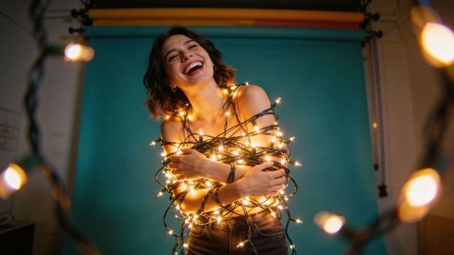 Joyful woman wrapped in glowing fairy lights laughing against teal backdrop, candid portrait