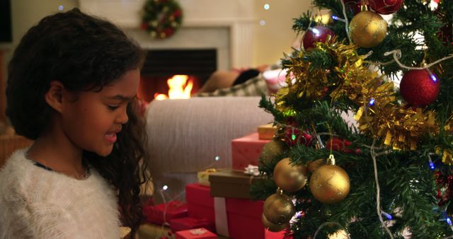 Biracial girl decorates a Christmas tree at home. She's immersed in the festive spirit, surrounded by gifts and warm fireplace glow.