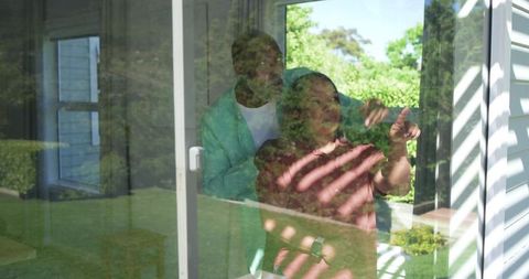 Diverse senior couple standing at glass door pointing to garden, sunlight through blinds