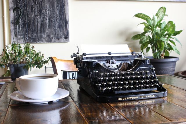 Vintage typewriter placed on wooden table next to coffee cup in a cozy café setting with green plants. Perfect for use in blog posts about writing, creativity, or retro lifestyle. Ideal for promoting coffee shops, creative spaces or freelance work environments.