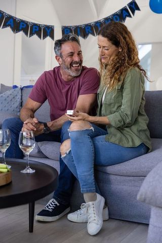 Mature couple celebrating birthday at home while opening gift and laughing on cozy sofa