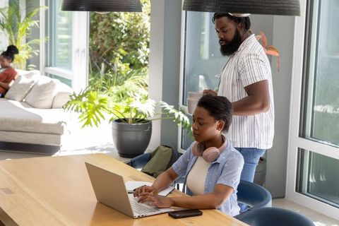 Young woman studying on laptop at sunlit modern openplan home table with headphones and family