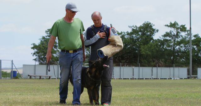 Professional dog trainers engaging in a training session with a German Shepherd in an outdoor grassy field. One trainer is wearing protective gear for handling the dog. Ideal for demonstrating dog training techniques, animal behavior training, and professional pet training scenarios.