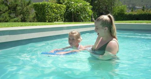 Young girl learning to swim with caregiver using blue kickboard in sunny backyard pool
