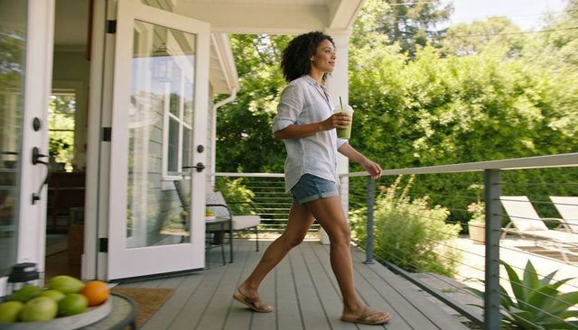 Woman walking on sunny deck holding green smoothie near open french doors and garden