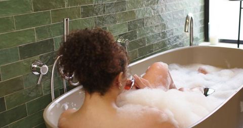 African american woman soaking in bubble bath holding wine glass by green tiled bathroom