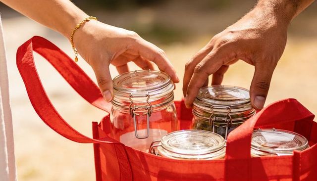 Hands placing glass jars into red reusable tote in warm sunlight