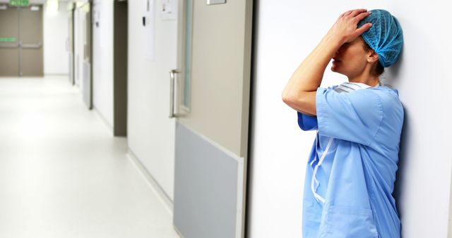 Stressed nurse leaning against wall in hospital corridor, capturing emotional exhaustion and burnout in healthcare setting. Useful for articles related to mental health in medical professions, healthcare stress, workplace pressure, and hospital working conditions. Can be used on blogs, news stories, and educational resources focusing on healthcare professionals' well-being.