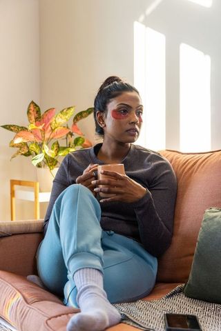 Indian woman relaxing on sofa with hydrogel eye patches holding mug in cozy sunlight