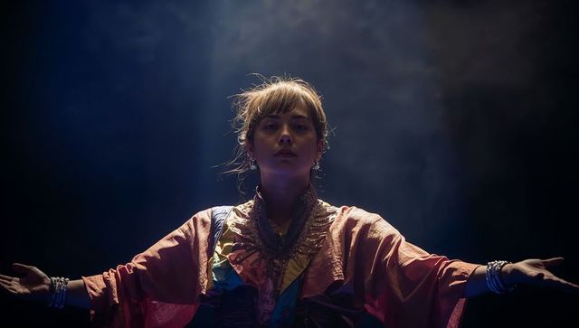 Dramatic stage performer extending arms in spotlight wearing layered robes and jewelry
