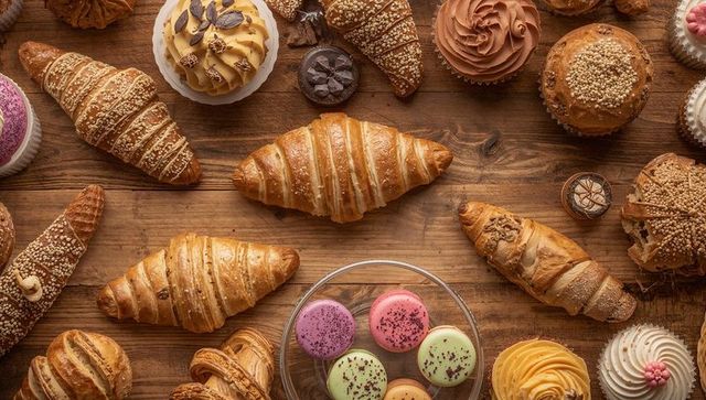 Golden croissant with colorful macarons, cupcakes and seeded rolls on rustic wood table