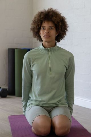 African-american woman kneeling on yoga mat wearing sage activewear in calm home studio