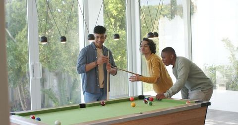 Diverse friends playing pool in modern home game room with glass wall and pendant lights