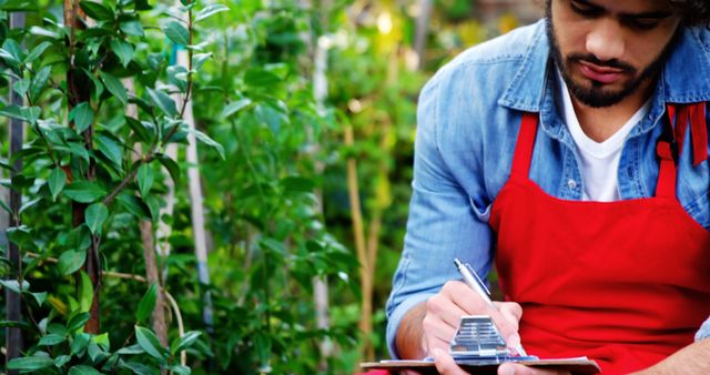 Young Man in Red Apron Taking Notes in Lush Garden - Download Free Stock Images Pikwizard.com