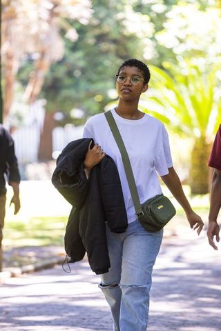 African american woman walking in park holding puffer jacket and green crossbody bag