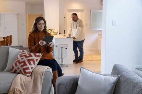 Middle-aged woman sitting on sofa holding tablet while partner leaning on kitchen counter in cozy co