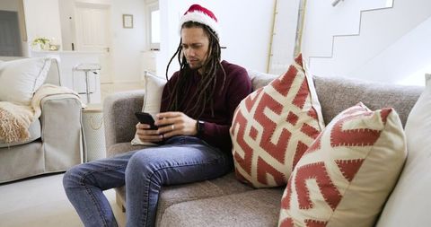 Dreadlocked man wearing santa hat sitting on sofa checking smartphone in cozy living room