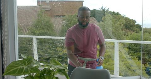 African man cleaning balcony glass door with squeegee and spray bottle during rainy day