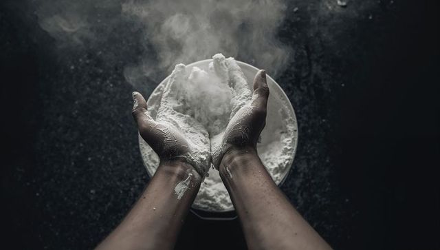 Hands sifting flour over bowl with rising dust on dark kitchen countertop