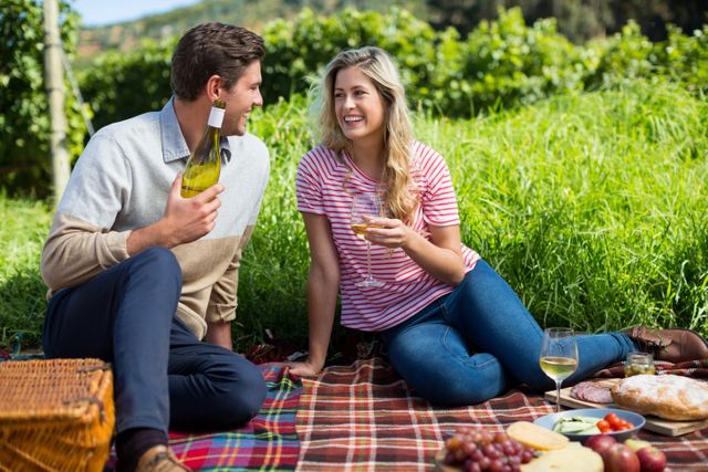 Couple sitting on picnic blanket in vineyard, holding wine bottle and glass, enjoying outdoor meal. Ideal for use in advertisements for wine, outdoor activities, romantic getaways, and lifestyle blogs.