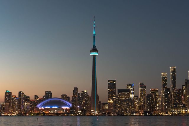 Skyline of Toronto at dusk, featuring the iconic CN Tower illuminated against a backdrop of vibrant city lights. The calm waters of the waterfront reflect the urban lights, creating a serene yet bustling ambiance. Ideal for use in travel brochures, promotional materials for tourism, websites highlighting Canadian cities, desktop wallpapers, and social media posts celebrating cityscapes.