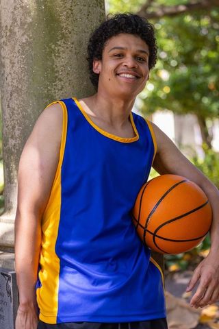 Smiling african american athlete leaning on tree holding basketball in blue yellow jersey