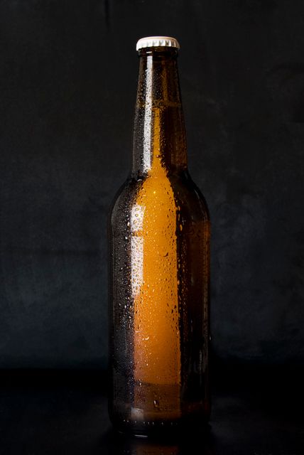 Chilled brown glass beer bottle with droplets of condensation standing against a dark background. Ideal for advertising cold beverages, alcoholic drinks, or for use in promotional materials for bars, pubs, and breweries. Can also be used in online articles discussing beer varieties or brewing processes.