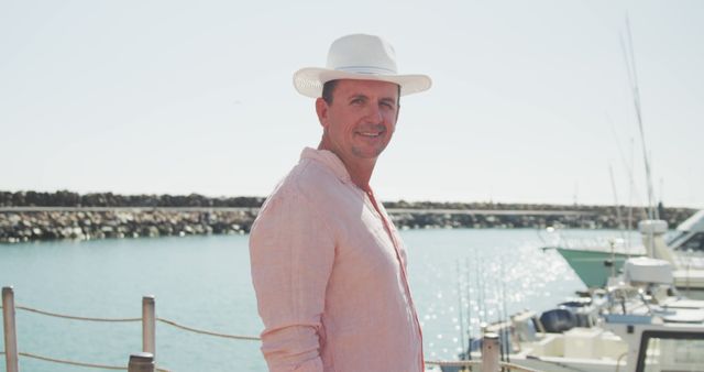 Portrait of smiling caucasian man in hat standing on jetty by boats in harbour on a sunny day. Leisure, free time, travel and vacations.