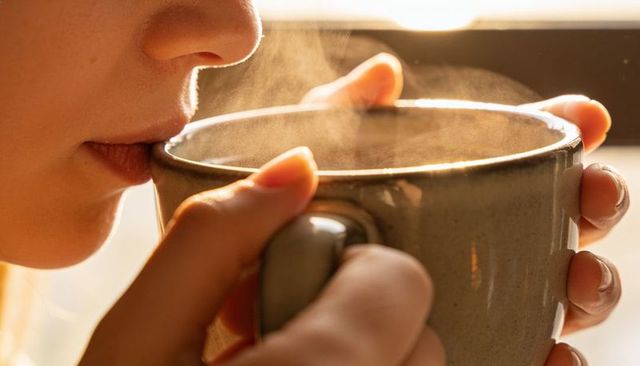 Holding steaming ceramic mug near window, sipping warm beverage closeup