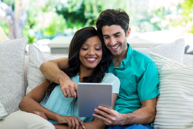 Young couple sitting on a comfortable sofa at home, using a digital tablet together. They are smiling and appear to be enjoying their time. This image can be used for promoting home technology, lifestyle blogs, relationship advice articles, or advertisements for home decor and furniture.