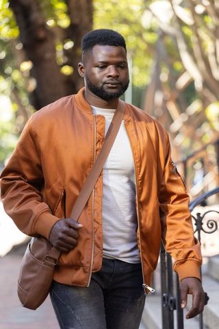 Man walking wearing rust-orange bomber jacket and brown crossbody bag on sunlit sidewalk