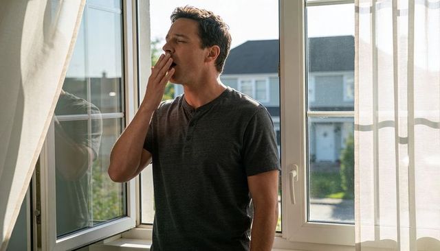 Yawning man covering mouth at open window in bright suburban morning light wearing henley shirt