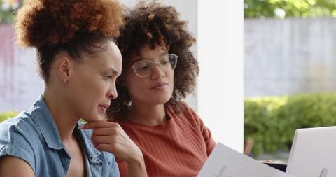 Two women collaborating over laptop and documents in sunlit casual workspace by window