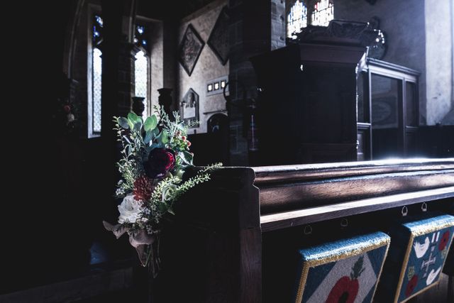Flower arrangement on a church pew in a dimly lit historic church creates a serene and atmospheric setting. Stained glass windows and wooden benches indicate a place of worship and their connection to the floral arrangement. Use in contexts related to churches, historical buildings, religious ceremonies, peaceful environments, or tranquil interior settings.