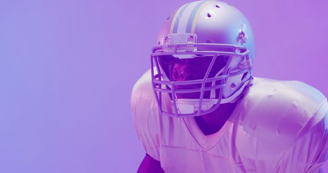 American football player wearing protective helmet and gear stands under dramatic purple lighting. Ideal for use in sports-related advertisements, posters promoting football events, fitness and health campaigns, and articles on athletic training and football insights.
