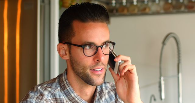 Young male professional wearing glasses in plaid shirt talking on phone in modern kitchen environment. Can be used for images depicting mobile communication, business-related discussions, remote work scenarios, or daily life in a home setting.