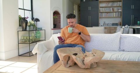 African american man sipping coffee and scrolling phone on sunlit modern sofa