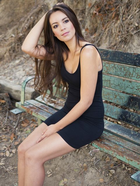 A young woman with long hair is sitting on a rustic green bench in a natural setting. She is wearing a casual black dress and appears to be relaxing. The scene suggests a tranquil moment in nature, with fallen autumn leaves around. This image can be used for lifestyle blogs, relaxation concepts, fashion promotions, or outdoor activity advertisements.