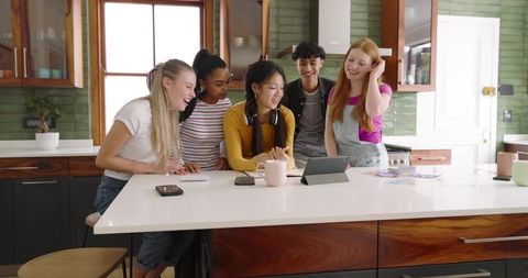 Diverse friends gathering around tablet on kitchen island, laughing and sharing ideas