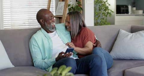 African american couple cuddling on sofa sharing smartphone and laughing in cozy home