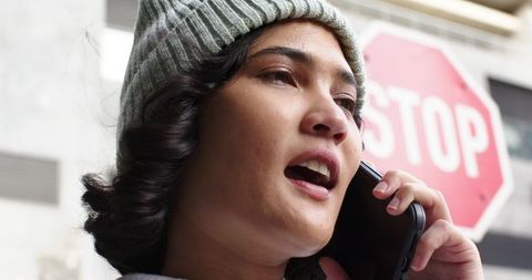 Woman speaking on smartphone wearing knit beanie near stop sign in urban street closeup